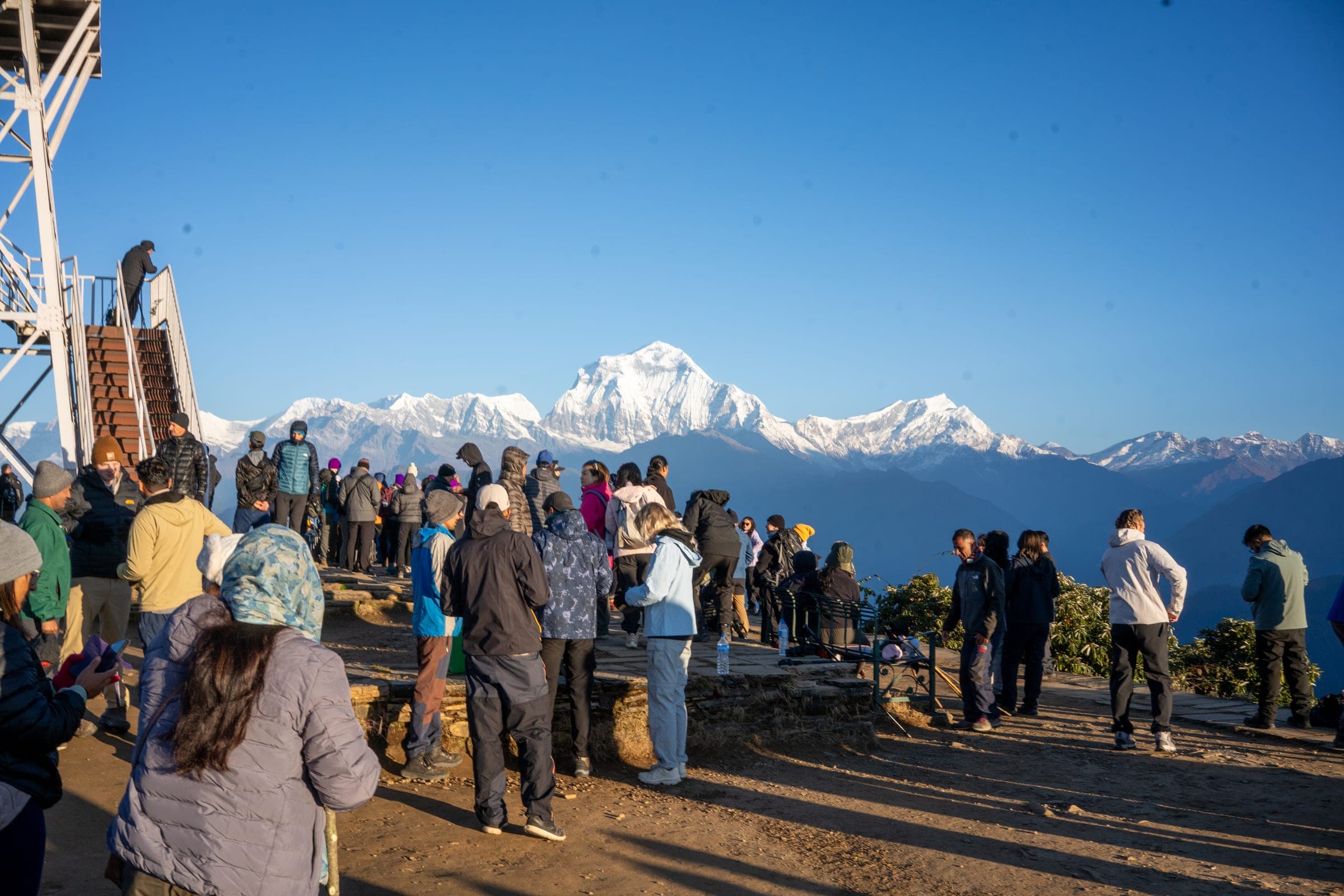 A crowd of trekkers on a rocky viewpoint facing a snow-capped mountain range at sunrise