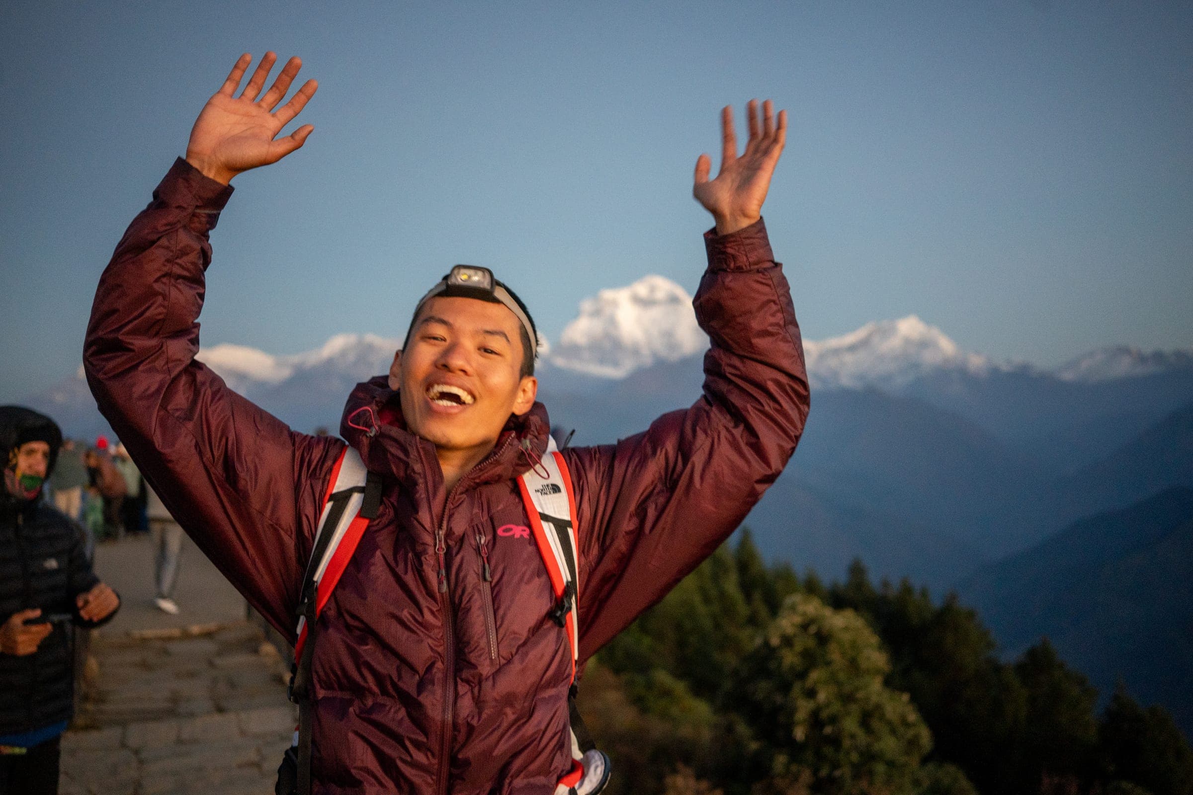 A hiker in an Outdoor Research jacket raising both arms at sunrise above snow peaks
