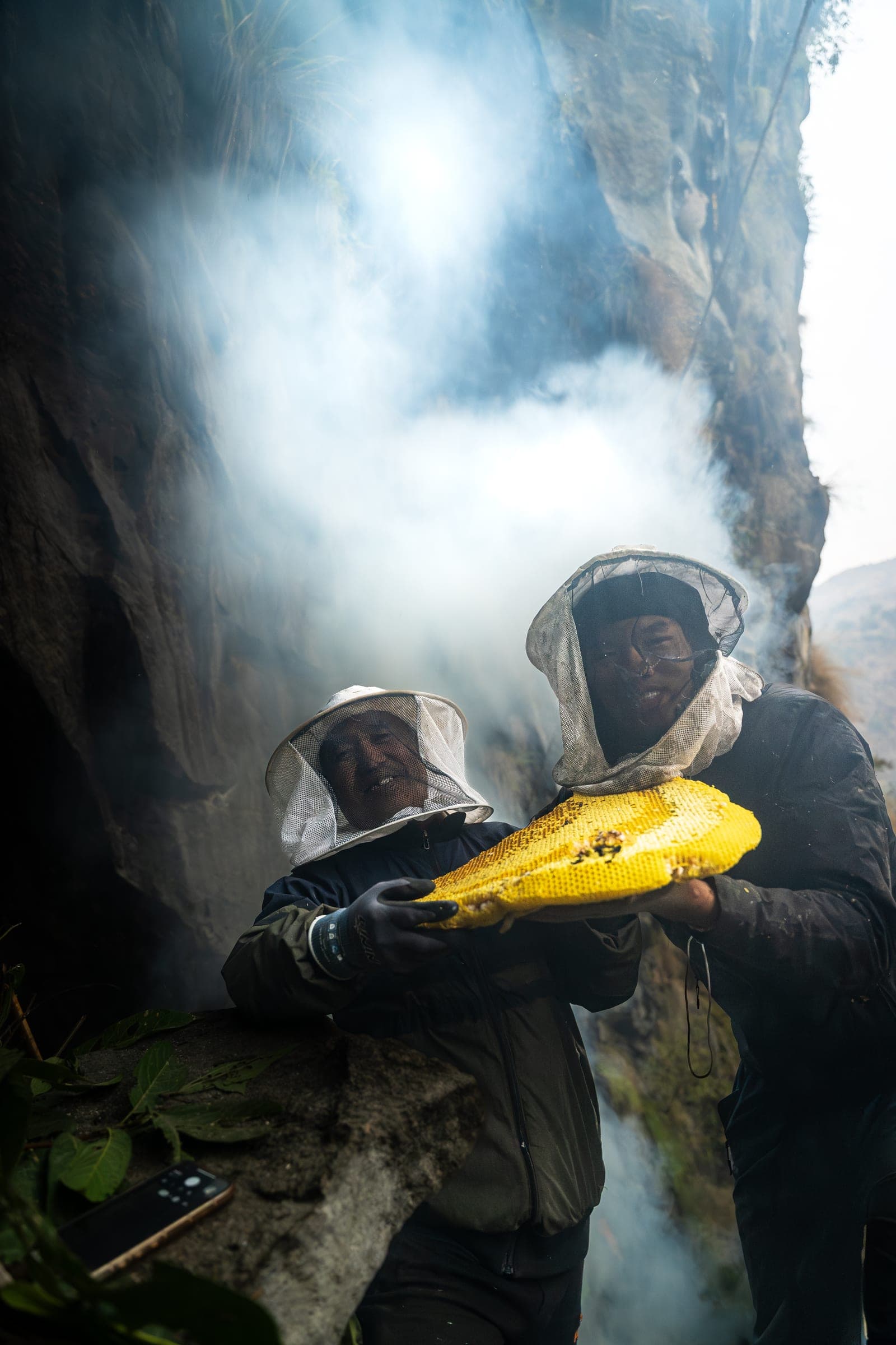 Two smiling honey hunters in white mesh veils holding a large yellow honeycomb with smoke behind them