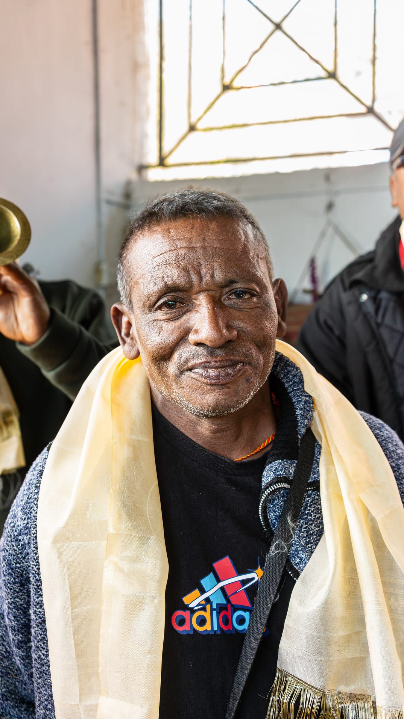 An older man in a cream shawl and knit cardigan smiling at the camera