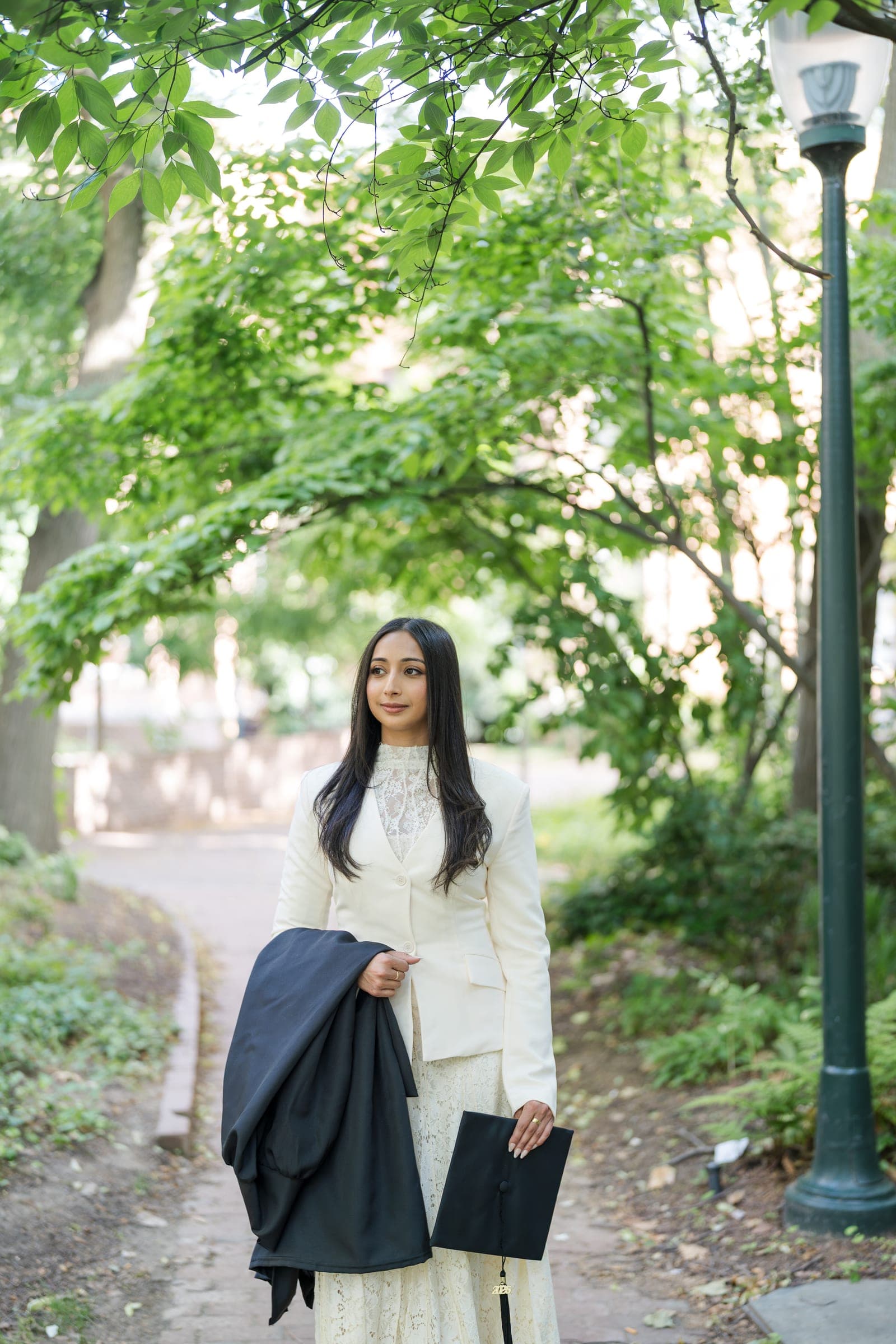 A woman in a white blazer holding a black graduation gown and cap on a brick path