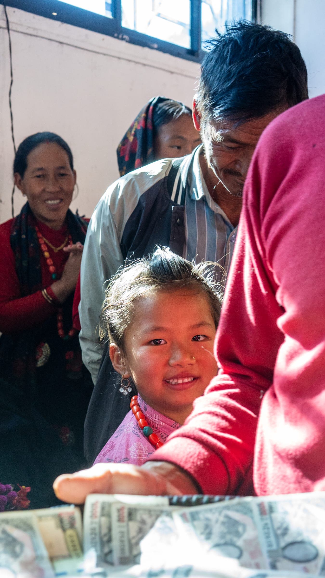 A young girl in a pink top and red-bead necklace smiling in a crowded marketplace