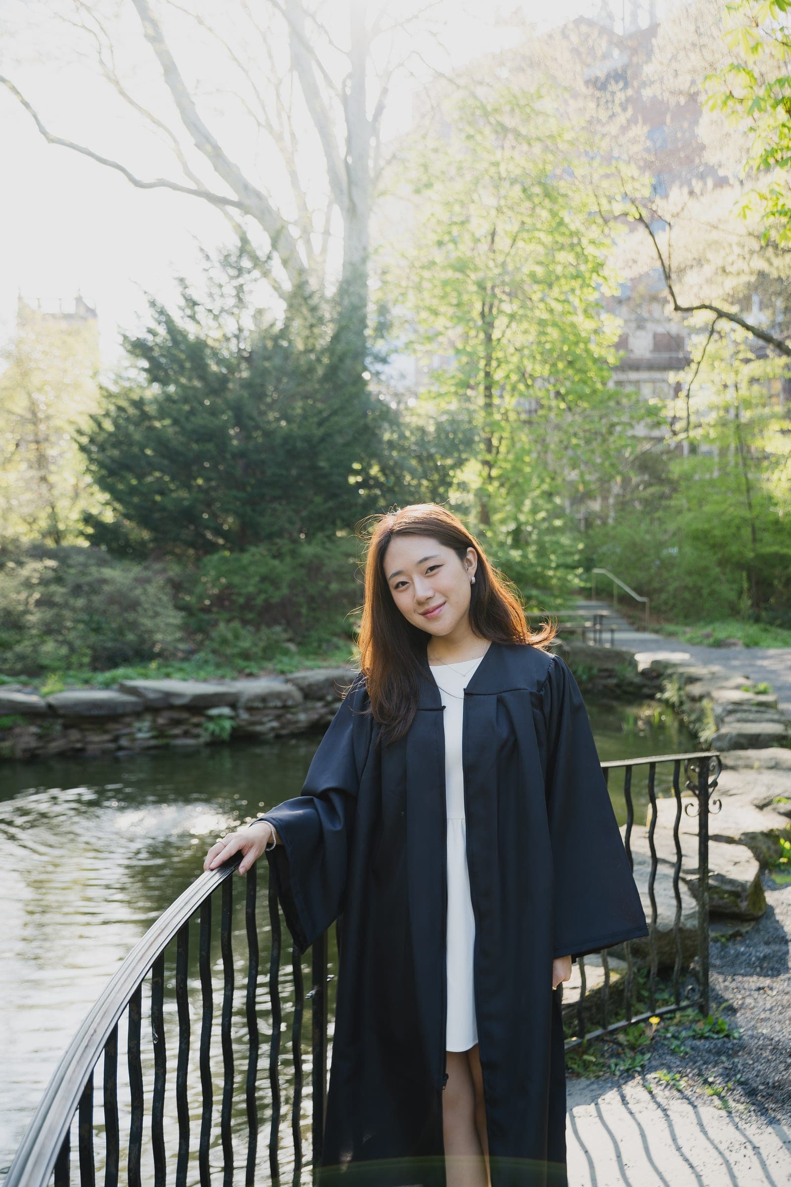 A graduate in an open black gown and white dress standing by a pond