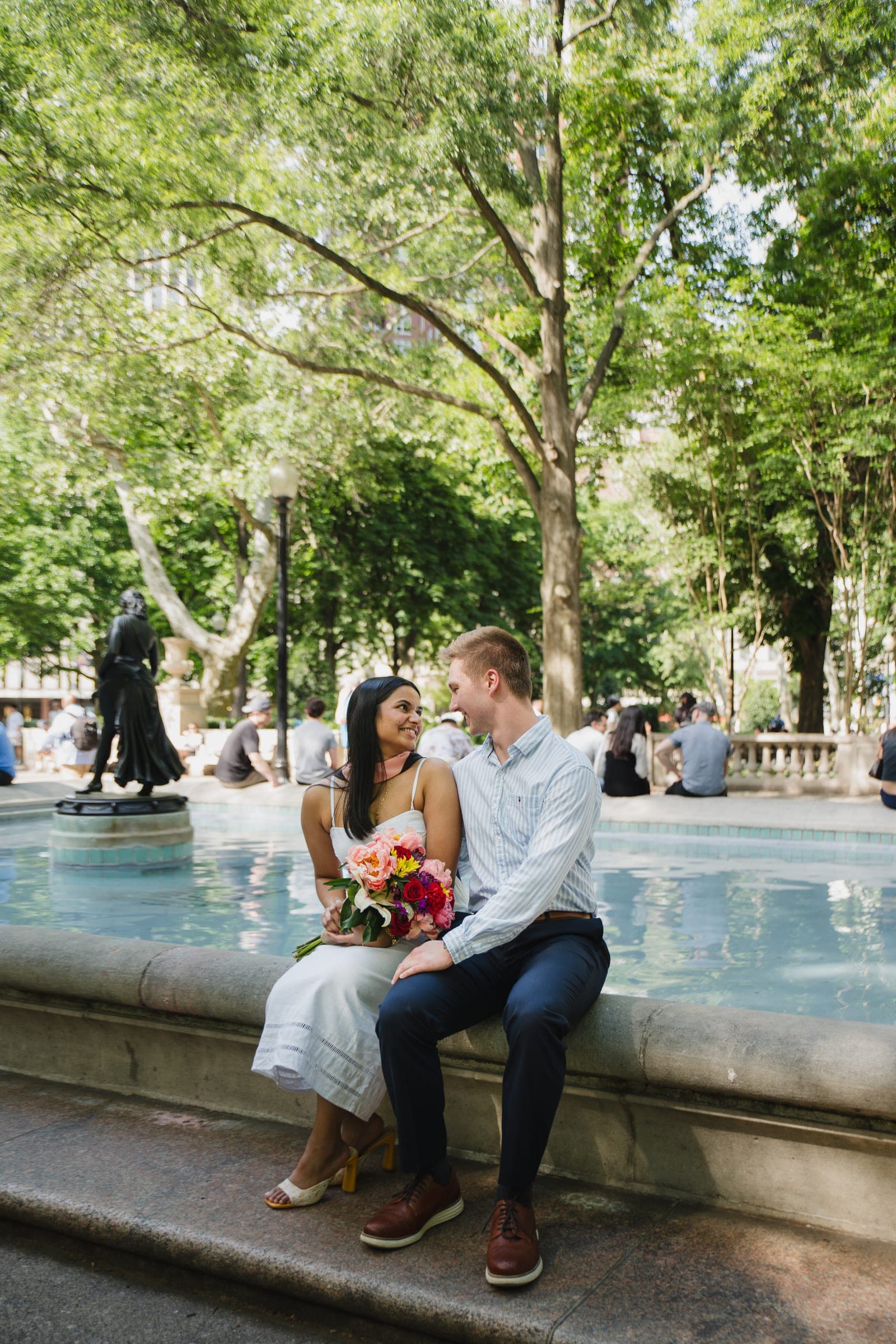 A couple sitting on a fountain rim in a city park, the woman holding a bright bouquet